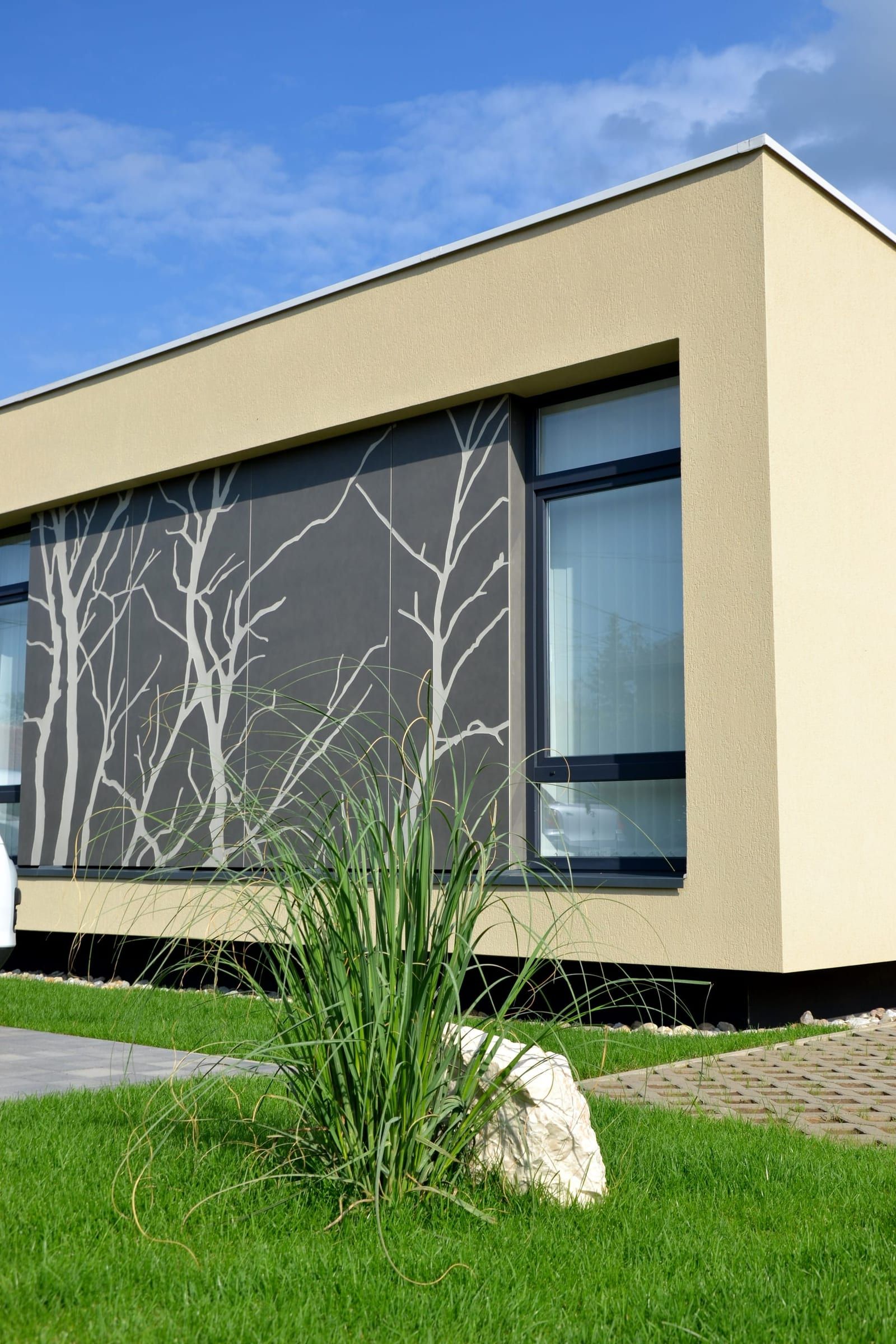Faágakat idéző mintázatú homlokzati panel az egészségházon, előtte pázsit és fűcsomó – L.Art Építészet